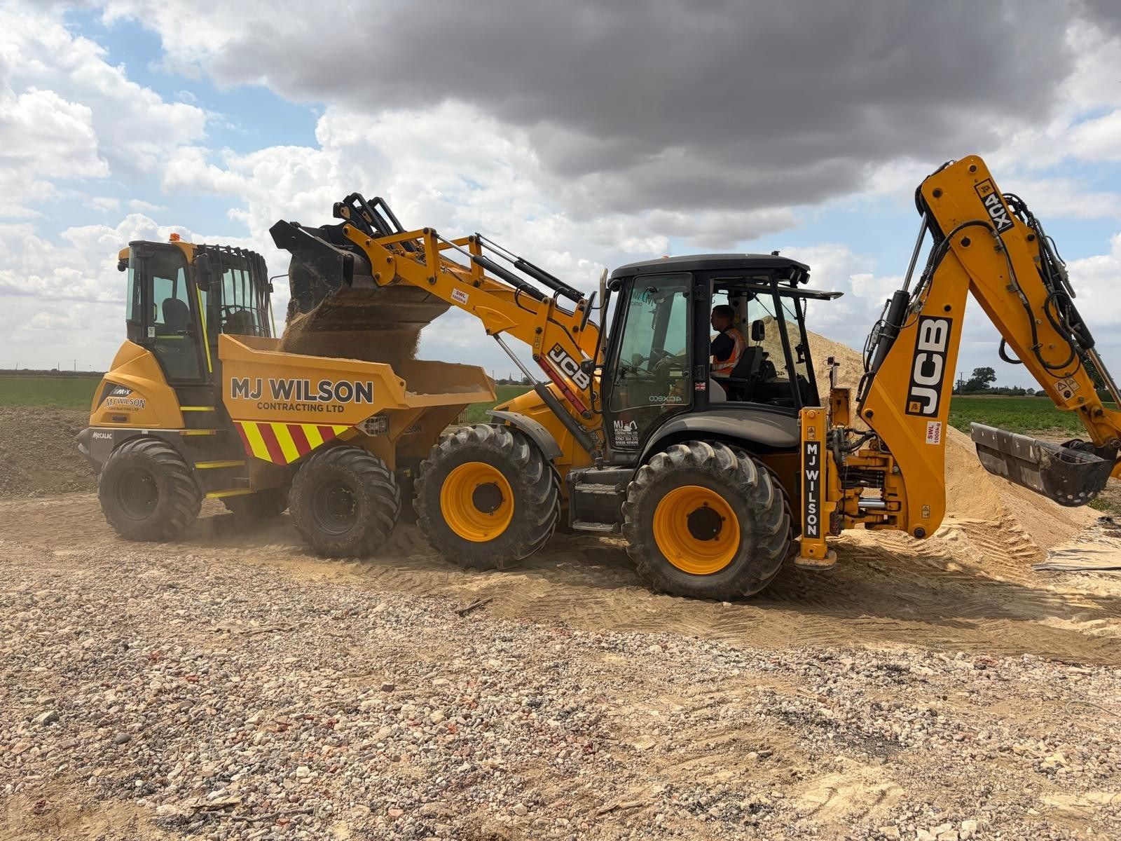 A JCB backhoe loader filling an M J Wilson dumper with sand on a site